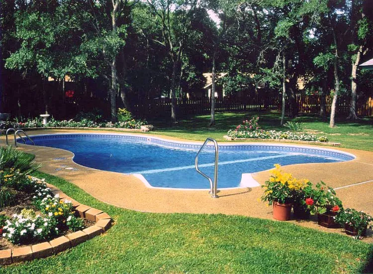 Figure-eight shaped pool with blue vinyl liner, concrete apron, and colorful flower garden beds in a wooded backyard in Lakehurst, NJ by Gorlin Pools