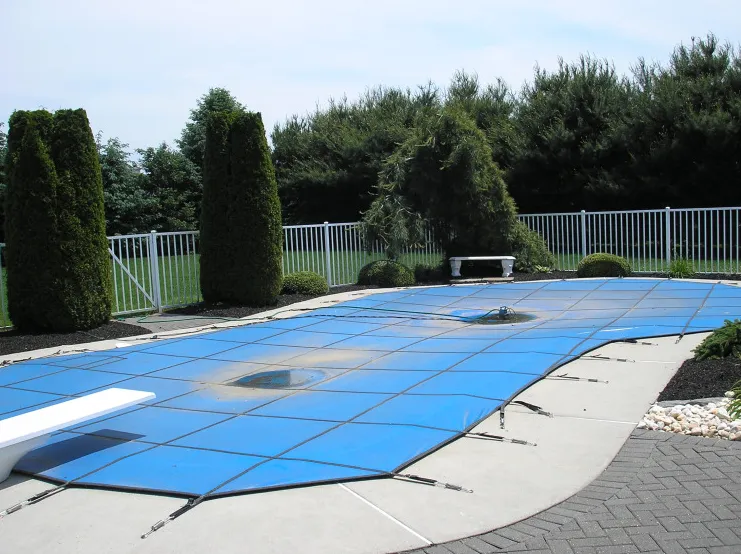 Freeform pool with blue mesh safety cover, diving board, and concrete deck bordered by arborvitae trees and white aluminum fence in Lakehurst, NJ by Gorlin Pools