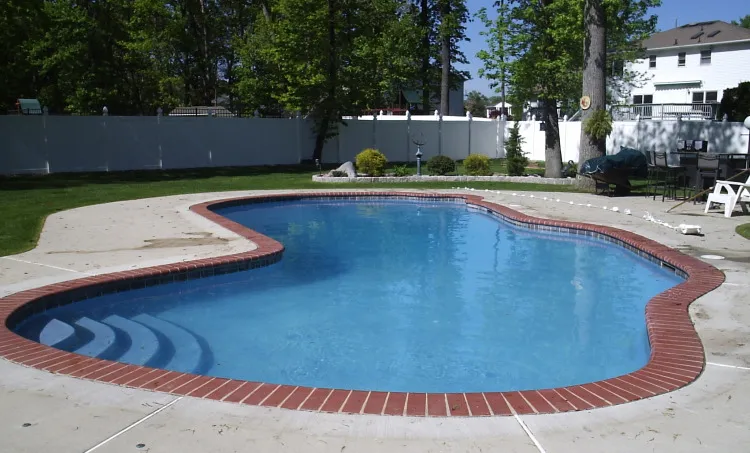 Freeform pool with red brick coping, built-in corner steps, and concrete deck surrounded by a white vinyl fence in Lakehurst, NJ by Gorlin Pools