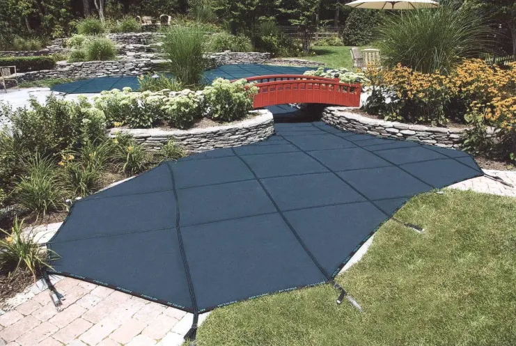 Freeform pool with dark mesh safety cover installed, decorative red arched bridge, and stone wall garden in Lakehurst, NJ by Gorlin Pools