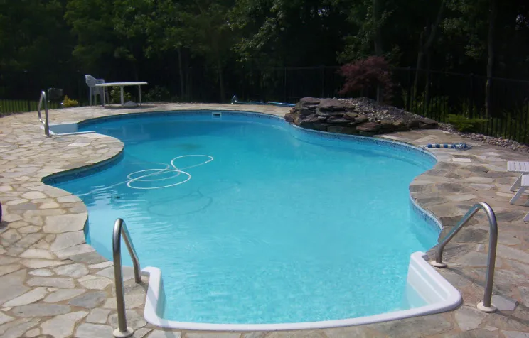 Freeform pool with dual stainless steel handrails, irregular flagstone patio, and natural rock feature in a wooded backyard in Lakehurst, NJ by Gorlin Pools