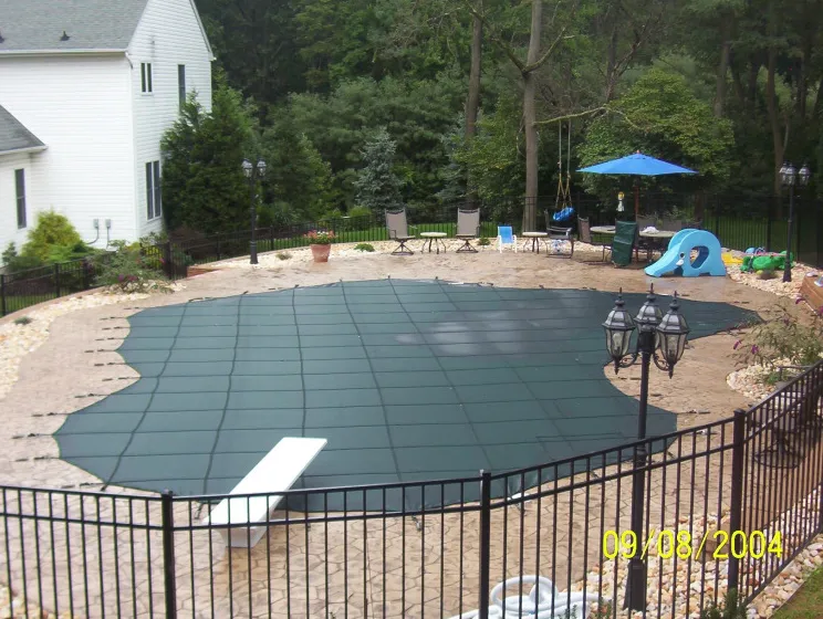 Freeform pool with green mesh safety cover and diving board on a flagstone patio with black iron fence and backyard play area in Lakehurst, NJ by Gorlin Pools