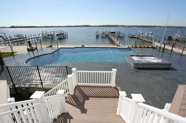 Geometric pool with glass tile spa, dark slate deck, and panoramic bay marina view seen from a white railing staircase in Lakehurst, NJ by Gorlin Pools