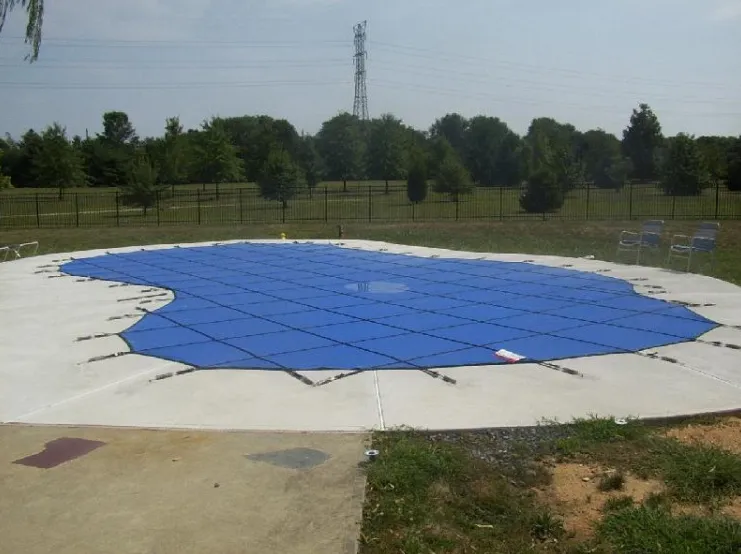 Large freeform pool with anchored blue mesh safety cover on a concrete deck in an open grassy yard with black iron fence in Lakehurst, NJ by Gorlin Pools