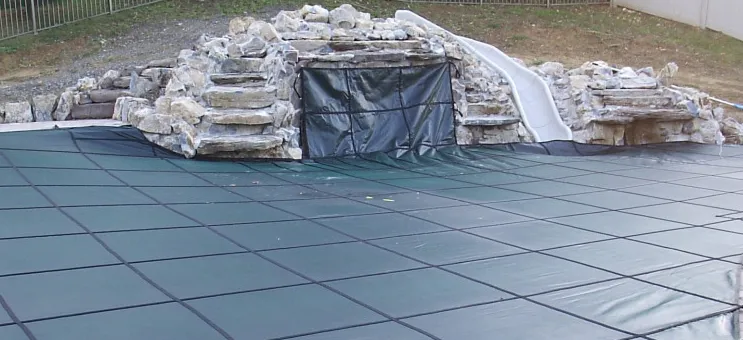 Green mesh safety cover installed over a pool with a rock waterfall grotto and white slide during winter closing in Lakehurst, NJ by Gorlin Pools