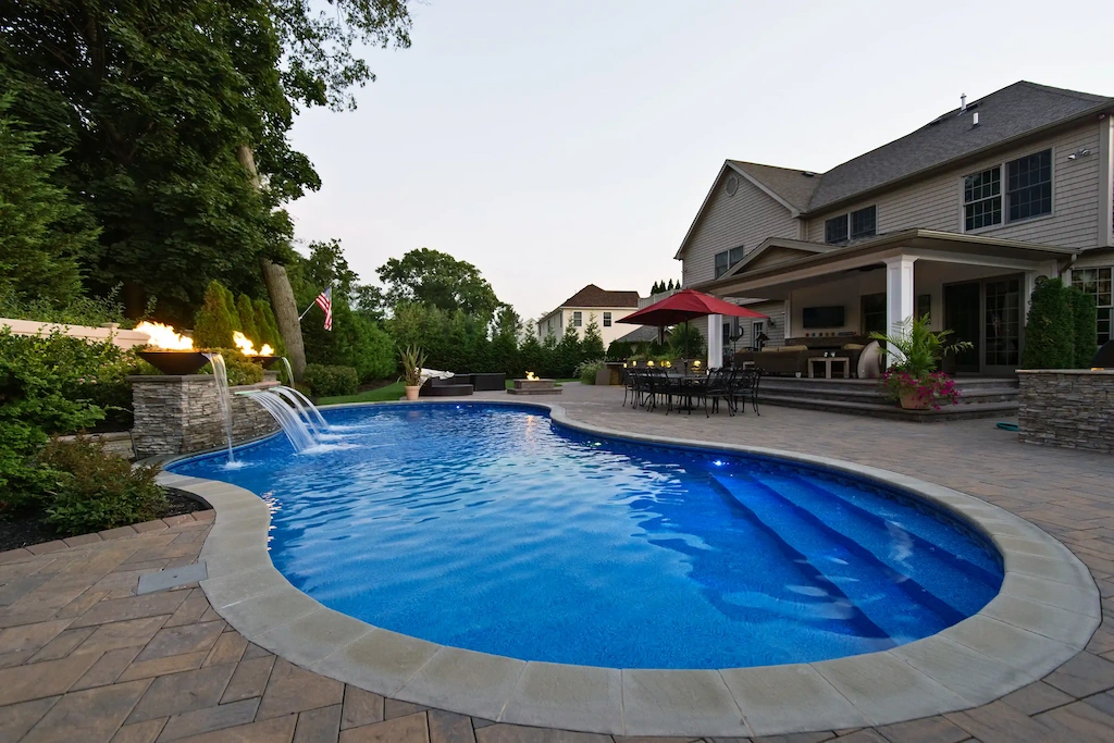 Freeform pool with fire bowl waterfalls, paver deck, and outdoor living space at dusk in Lakehurst, NJ by Gorlin Pools