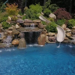 Custom pool with tiered rock waterfall, built-in spa, and water slide in a landscaped backyard in New Jersey by Gorlin Pools