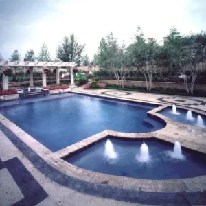 Formal geometric pool with water jet fountains, decorative mosaic tile border, and white pergola in a landscaped backyard in Lakehurst, NJ by Gorlin Pools