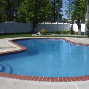 Freeform pool with red brick coping, built-in corner steps, and concrete deck surrounded by a white vinyl fence in Lakehurst, NJ by Gorlin Pools