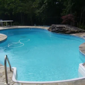 Freeform pool with dual stainless steel handrails, irregular flagstone patio, and natural rock feature in a wooded backyard in Lakehurst, NJ by Gorlin Pools