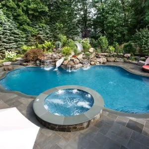 Freeform pool with raised circular spa, stone paver patio, rock waterfall, and slide surrounded by lush landscaping in New Jersey by Gorlin Pools