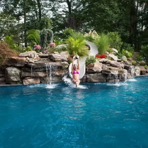 Child sliding into a freeform pool with rock waterfall feature and lush backyard landscaping in New Jersey by Gorlin Pools