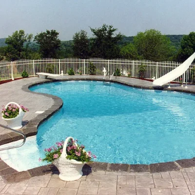 Kidney-shaped pool with diving board, white water slide, stamped concrete patio, and rolling hillside view in Lakehurst, NJ by Gorlin Pools