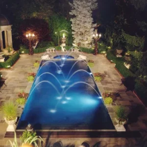 Aerial night view of a rectangular pool with arc water jet fountains, underwater lighting, and formal garden statuary in Lakehurst, NJ by Gorlin Pools