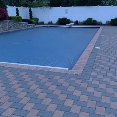 Rectangular pool with solid blue winter cover, brick coping, paver patio, and white vinyl fence at dusk in Lakehurst, NJ by Gorlin Pools