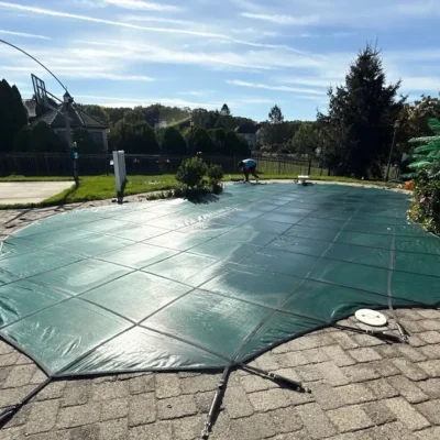 Technician securing green mesh safety cover with anchor straps over inground pool for winter closing – Gorlin Pools NJ